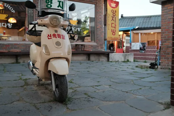 A cream delivery scooter parked on uneven stone paving outside small shops