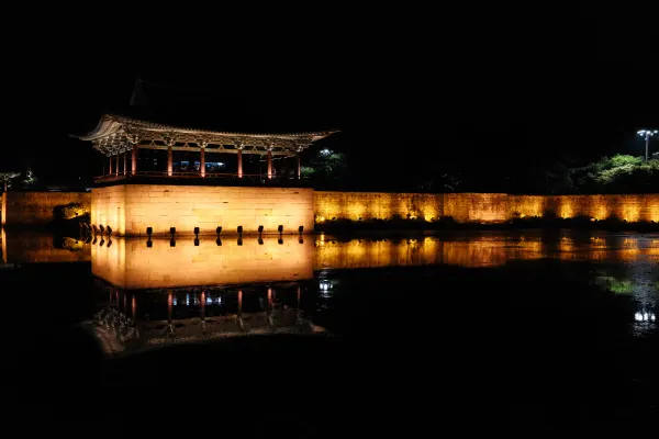 A traditional pavilion and wall glowing gold above their reflection at night