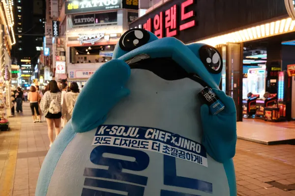 A blue Jinro soju mascot standing beneath bright shop signs on a busy night street