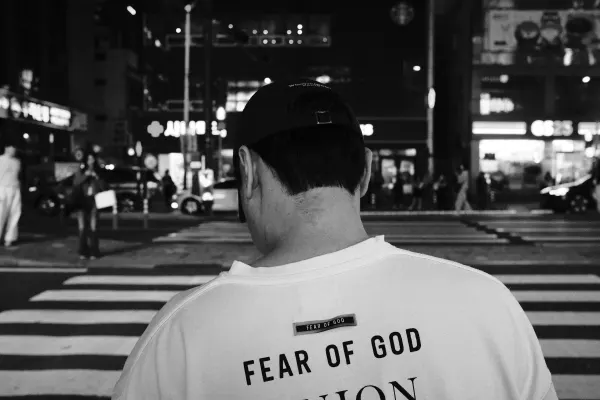 A person in a black cap waiting at a busy night crossing, photographed in black and white