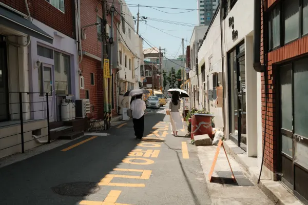 Pedestrians carrying umbrellas along a narrow sunlit lane