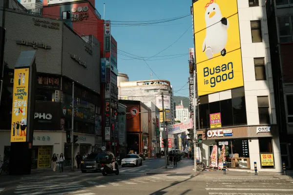 A sunlit city street beneath a yellow Busan is good sign