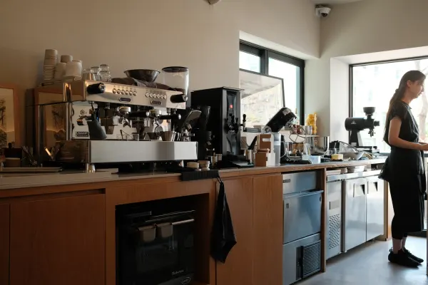 A sunlit cafe counter with an espresso machine and a person standing by the window